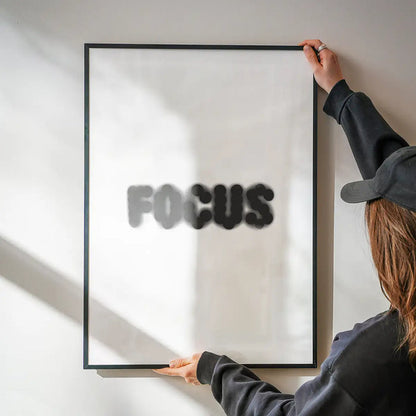 Person hanging a framed "FOCUS" quote print with blurred black text on white archival art paper.