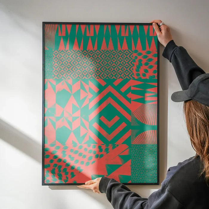 Hands hanging a framed pink and green abstract geometric print on a white wall.