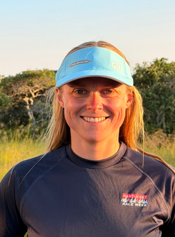 A smiling woman outdoors in a grassy field at sunset, wearing a light blue visor and a dark navy long-sleeve athletic shirt with a small red and white logo on the left chest.