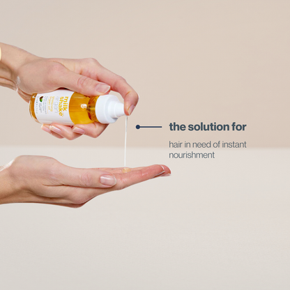 Hand holding a bottle of milk_shake hair oil with a droplet being released onto another hand against a beige background.