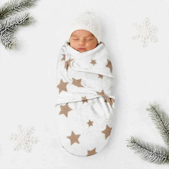 A sleeping newborn baby swaddled in a white fleece blanket with brown star patterns, wearing a matching white knit beanie, laid flat on a white background with decorative snowflakes and pine branches.