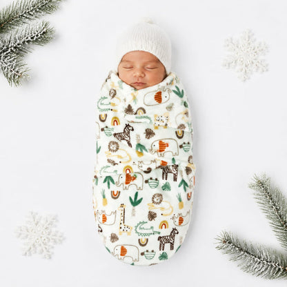 A sleeping newborn baby wrapped in a cream-colored swaddle blanket with a playful safari animal print, wearing a matching cream knit beanie. The scene is styled with white snowflakes and pine branches on a clean white background.