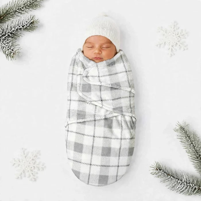 A sleeping newborn baby swaddled in a soft gray and white plaid blanket, wrapped in a white knit hat, laid on a white background with decorative snowflakes and pine branches.