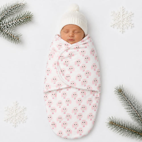 A sleeping newborn baby swaddled in a white blanket with a pink sheep pattern, wearing a white knit beanie hat, styled on a white background with decorative snowflakes and pine branches.