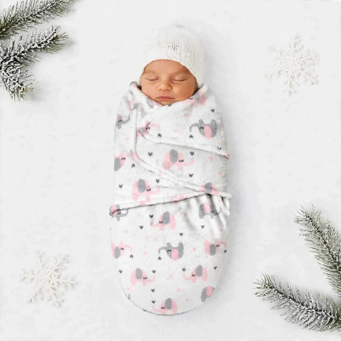 A sleeping newborn baby swaddled in a soft white blanket with a pink and gray elephant print, wearing a white knit hat, laid on a white background with decorative pine branches and snowflakes.
