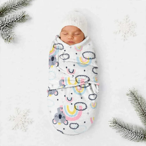 A sleeping newborn baby wrapped in a white swaddle blanket with a colorful cartoon rainbow and cloud pattern, wearing a white knit hat, styled on a white background with snowflake decorations and pine branches.
