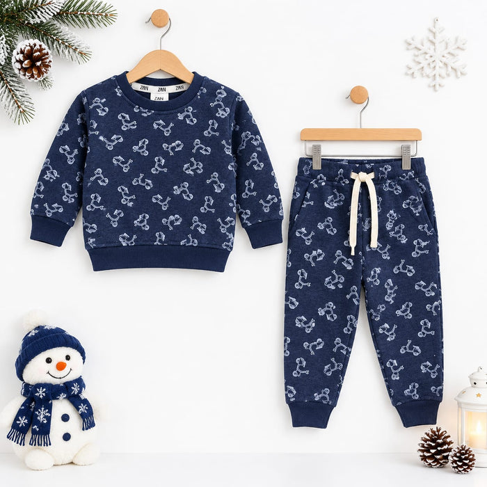 A children's two-piece pajama set hanging on wooden hangers against a white wall, featuring a navy blue sweatshirt and matching joggers with a white bone pattern. The set is styled with winter props including a snowman plush, pinecones, a snowflake orname
