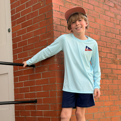 Young boy standing on steps against a brick wall wearing a light blue long-sleeve shirt and dark shorts.