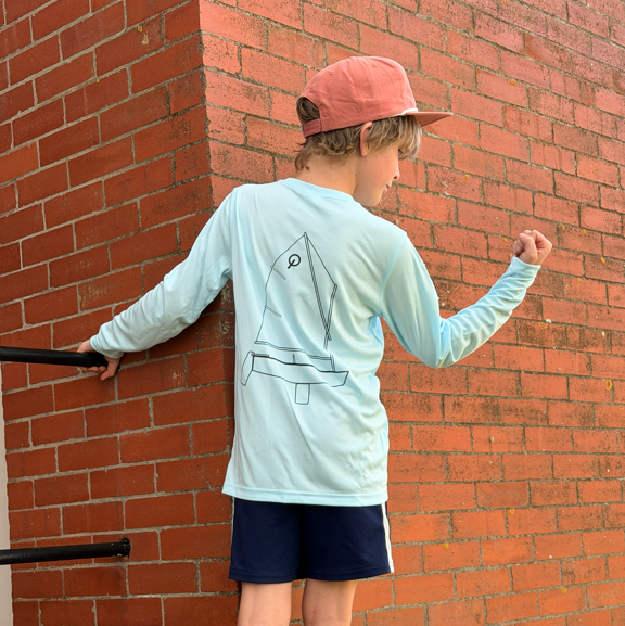 Child wearing a light blue long-sleeve shirt with a graphic design, navy shorts, and gray shoes against a brick wall.