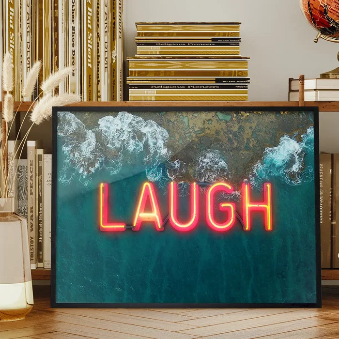 Framed wall art featuring a vibrant red neon LAUGH sign layered over an aerial ocean wave print on glossy paper.