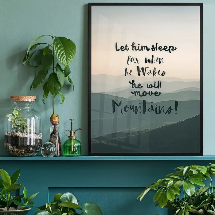 Let Him Sleep" quote print with subtle mountain background, framed in black, on a teal shelf.