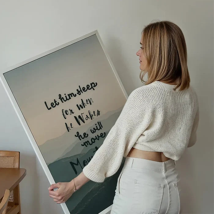 Woman holding a framed "Let Him Sleep" quote print with subtle mountain background and Scandinavian style.