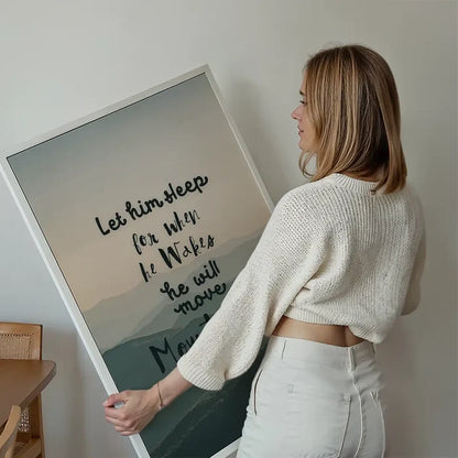 Woman holding a framed "Let Him Sleep" quote print with subtle mountain background and Scandinavian style.