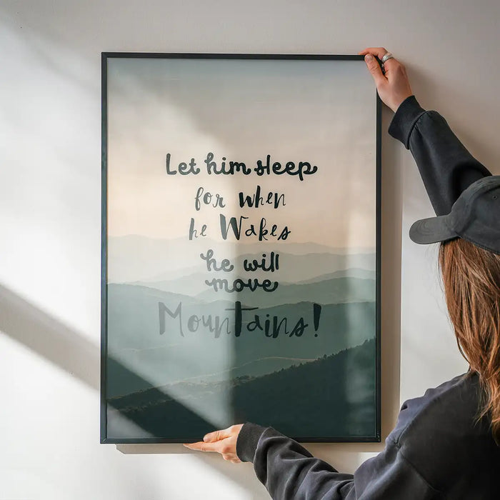 Let Him Sleep" quote print with subtle mountain background, held by hands against a white wall.