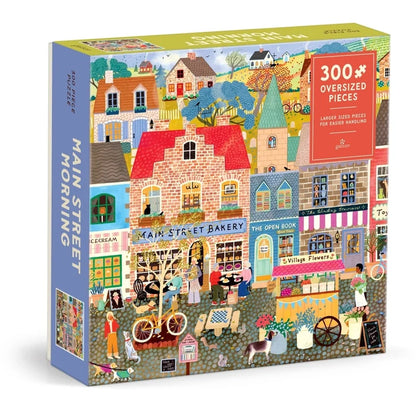 The Galison Main Street Morning 300 Piece Jigsaw Puzzle features a cozy and charming illustration of bustling shops, friendly people, and playful animals along a lively town street. The puzzle includes 300 oversized pieces for easy handling.