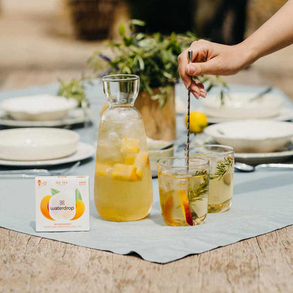 Person stirring a glass of iced tea with lemon and peach slices on a table with 'waterdrop' tea packaging.