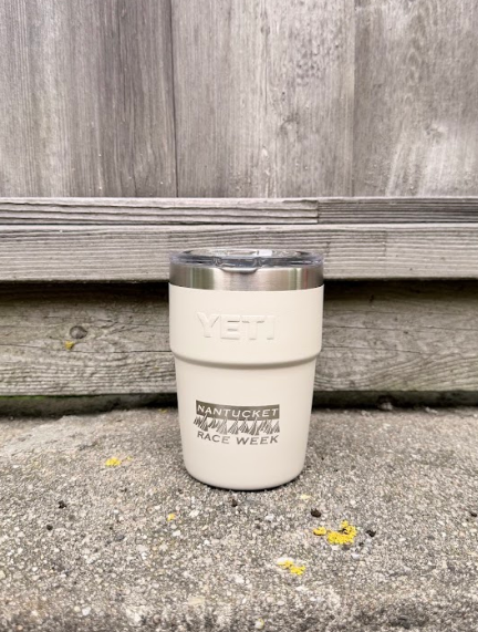A cream-colored YETI tumbler with a stainless steel lid sits on a concrete ledge in front of a weathered gray wooden fence.
