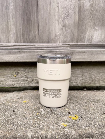A cream-colored YETI tumbler with a stainless steel lid sits on a concrete ledge in front of a weathered gray wooden fence.