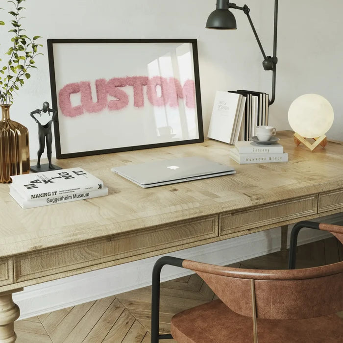Pastel pink fur-style "CUSTOM" name print in a black frame on a wooden desk with a laptop and decor.