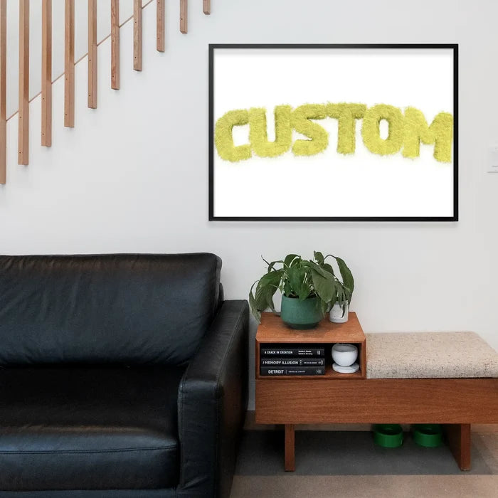 Pastel yellow fur style personalized name print in a black frame above a black leather sofa.