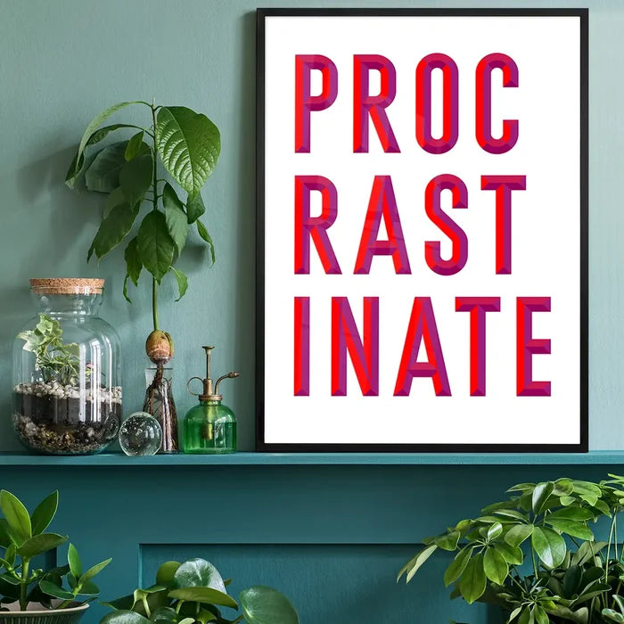 Black-framed "PROCRASTINATE" quote print with bold red and purple lettering on white archival paper, displayed on a teal shelf.