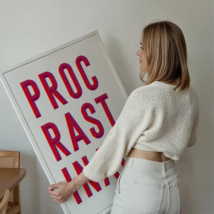 Woman holding a large white framed "PROCRASTINATE" typographic print with bold red and purple lettering.