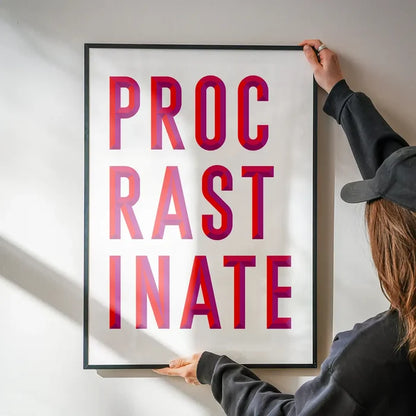Framed "PROCRASTINATE" quote print with bold red and purple lettering on white archival art paper.