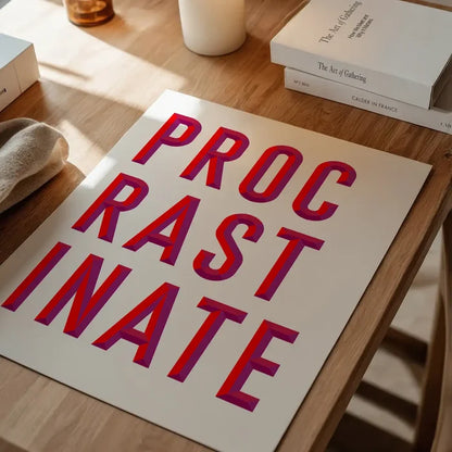 White art paper print with bold red and purple "PROCRASTINATE" typography on a wooden table.