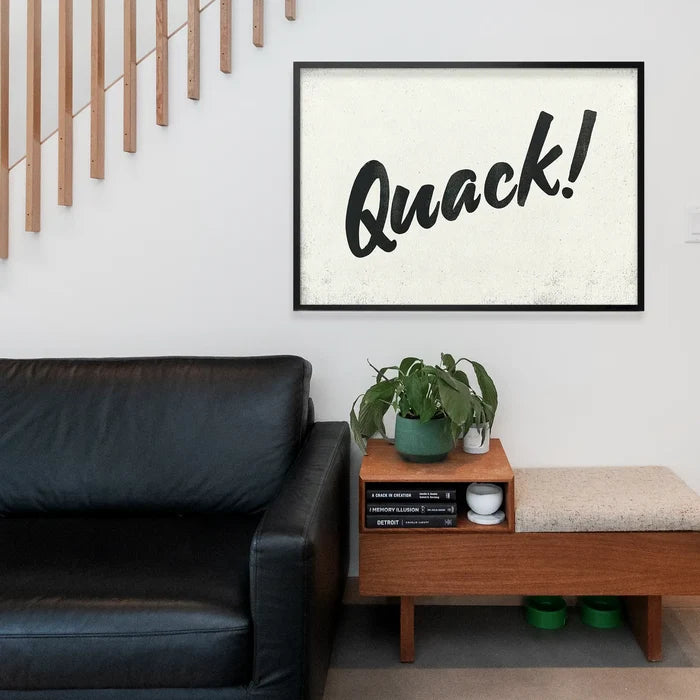 Black framed "Quack!" print with hand-painted brush script typography, displayed above a wooden bench and black leather sofa.