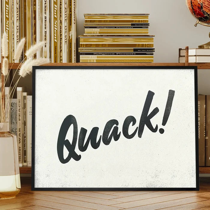 Framed "Quack!" animal sound print with black brush script on a textured white background, displayed on a wooden shelf.