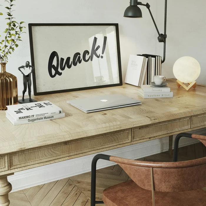 Minimalist "Quack!" animal sound print in a black frame on a light wood desk with books and a moon lamp.