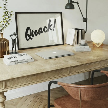 Minimalist "Quack!" animal sound print in a black frame on a light wood desk with books and a moon lamp.