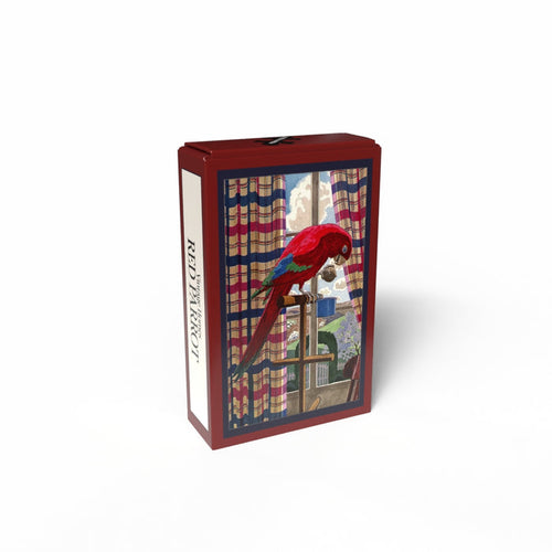 The Penny Puzzle "Red Parrot" is a 99 piece jigsaw puzzle in a rectangular box featuring a vintage-style illustration of a red parrot perched indoors by a window with striped curtains and trees visible outside.