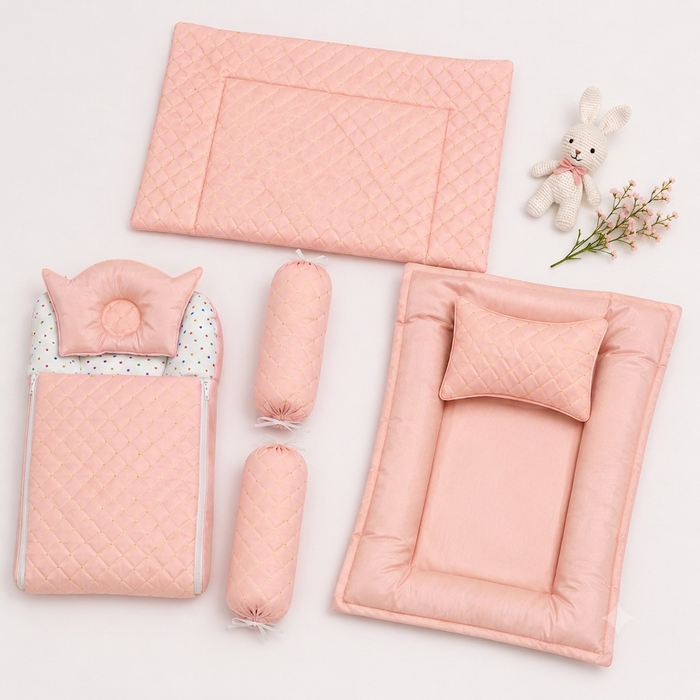 A coordinated baby bedding set arranged on a white background, including a large quilted pink mat, a smaller pink mat with a pillow, a matching folded pad, two cylindrical bolster pillows, a patterned insert, and a small stuffed bunny decoration.