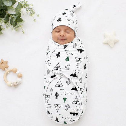 A sleeping newborn baby swaddled in a white muslin swaddle blanket with a black and green geometric teepee and cactus pattern, wearing a matching beanie. The baby is photographed on a white fabric background with baby props including a wooden star teether