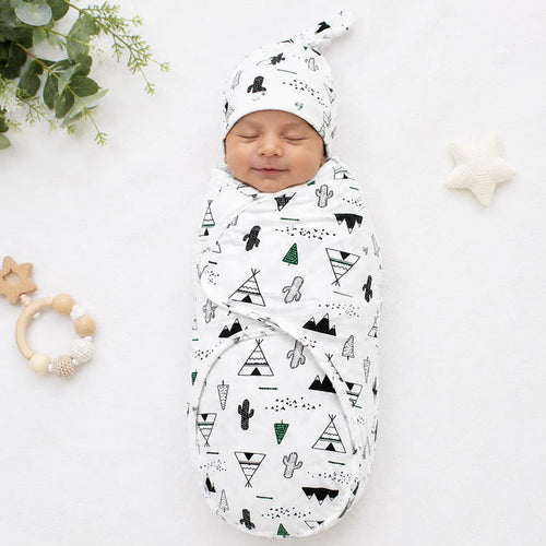 A sleeping newborn baby swaddled in a white muslin swaddle blanket with a black and green geometric teepee and cactus pattern, wearing a matching beanie. The baby is photographed on a white fabric background with baby props including a wooden star teether