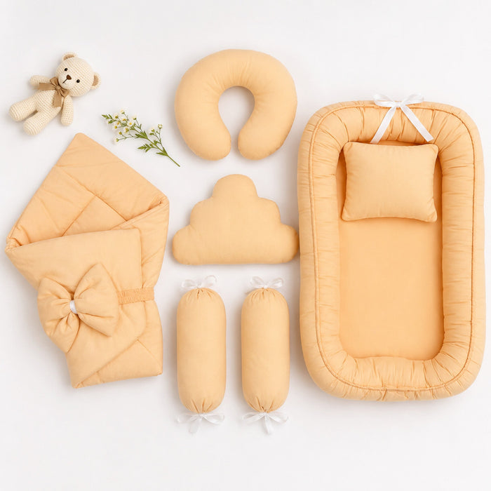 A coordinated baby bedding set arranged on a white background, including a bassinet with matching pillow, a folded blanket with bow, a U-shaped pillow, a cloud-shaped pillow, two cylindrical inserts, a small teddy bear prop, and a sprig of flowers.