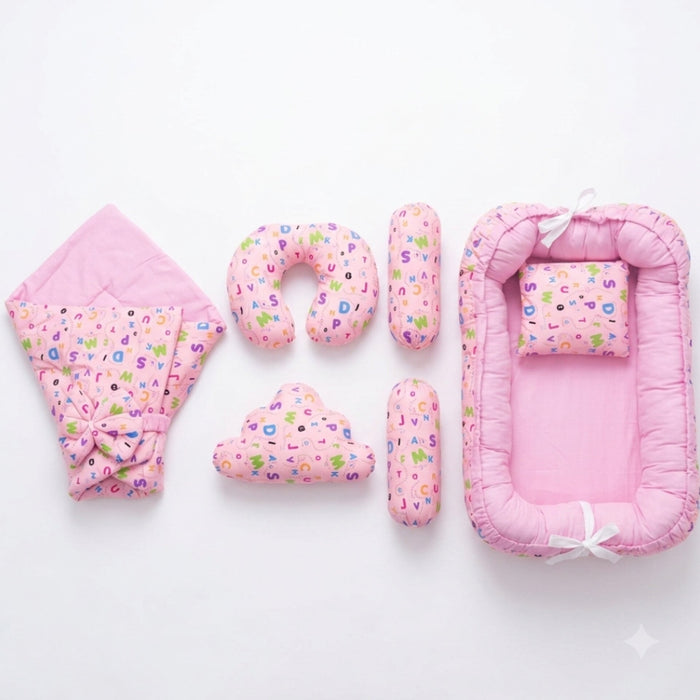 A pink baby bedding set arranged on a white background, including a bassinet, a hooded blanket, a U-shaped pillow, two cylindrical support pillows, and a cloud-shaped pillow. The fabric features a light pink base with a colorful alphabet pattern.