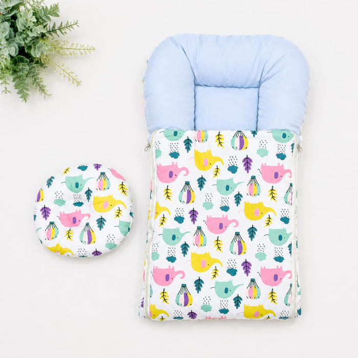 A baby footmuff and matching circular cushion set with a colorful pattern of pink teapots, yellow elephants, and green leaves on a white background, styled on a white surface with a green plant in the corner.