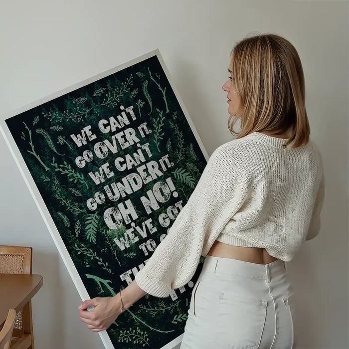 Blonde woman holding a large green typography print with "We Can't Go Over It" quote and botanical illustrations.