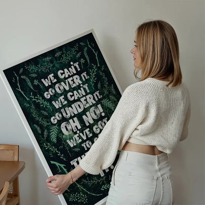 Blonde woman holding a large green typography print with "We Can't Go Over It" quote and botanical illustrations.