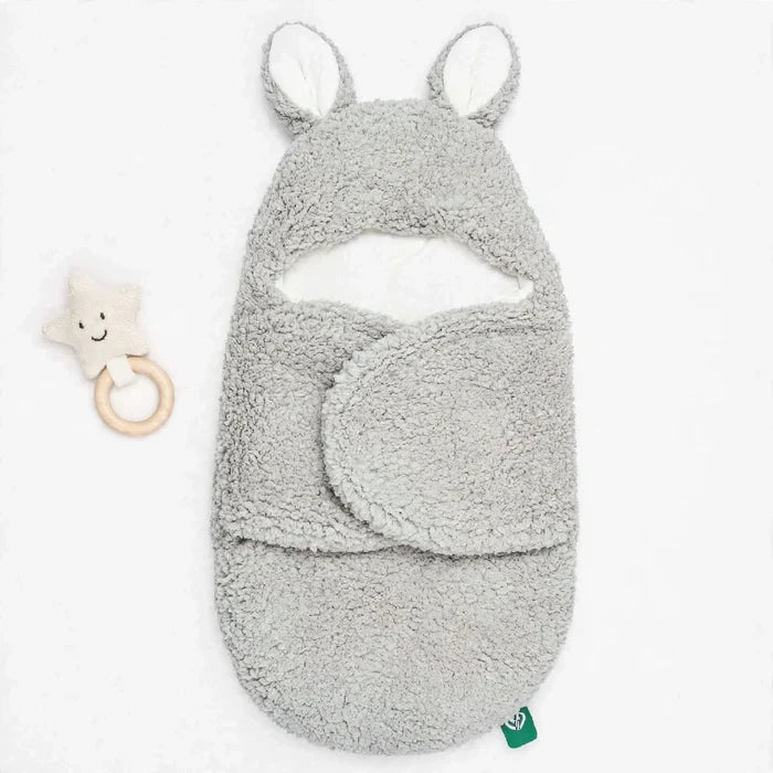 A light gray plush baby swaddle blanket with bunny ears and a matching footed cowl, shown on a white background next to a small star-shaped wooden teething toy.