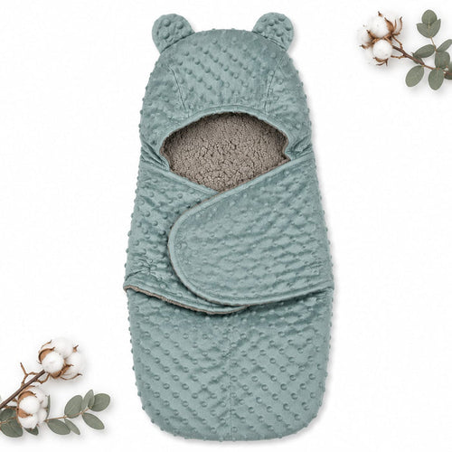 A baby swaddle wrap with a hood featuring bear ears, made of textured dusty blue minky fabric with a soft gray fleece lining. The swaddle is displayed flat on a white background with decorative cotton and eucalyptus sprigs in the corners.